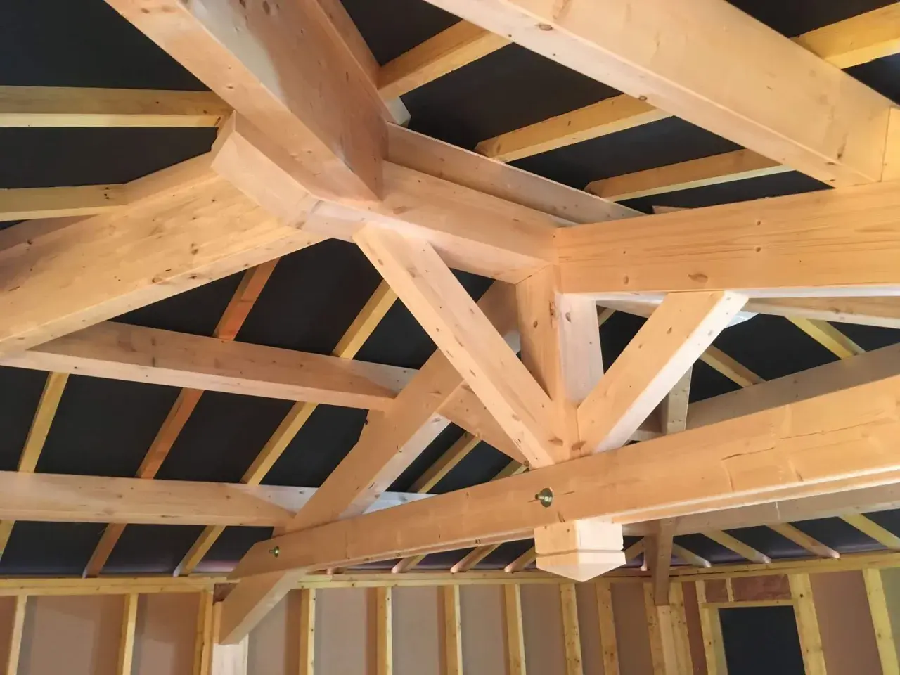Vue rapprochée d'une charpente traditionnelle 2 pans en bois clair, avec des poutres massives et des chevrons assemblés.