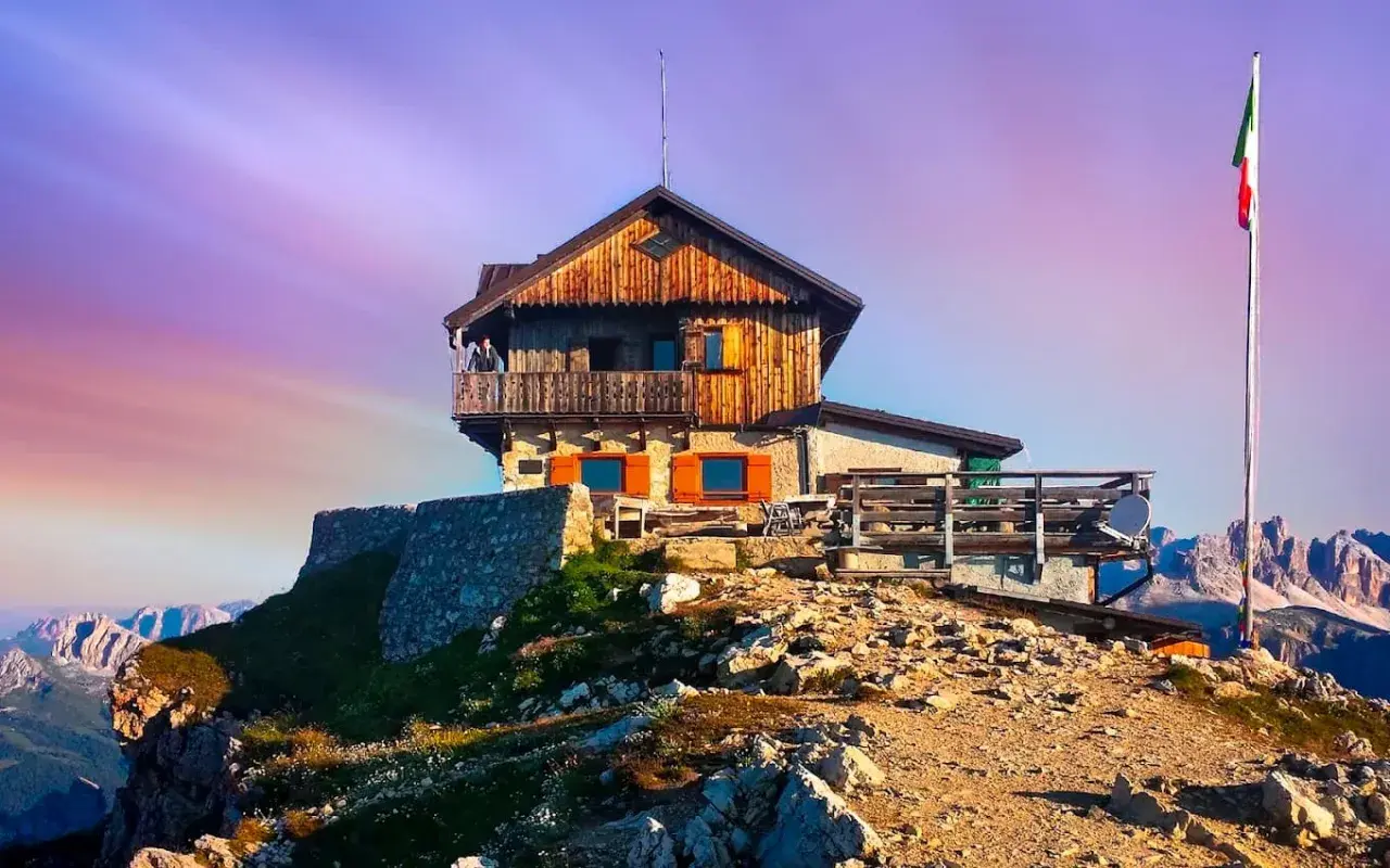 Rifugio Nuvolau thront auf einem Felsvorsprung unter einem lila Himmel. Eine italienische Flagge weht im Wind.