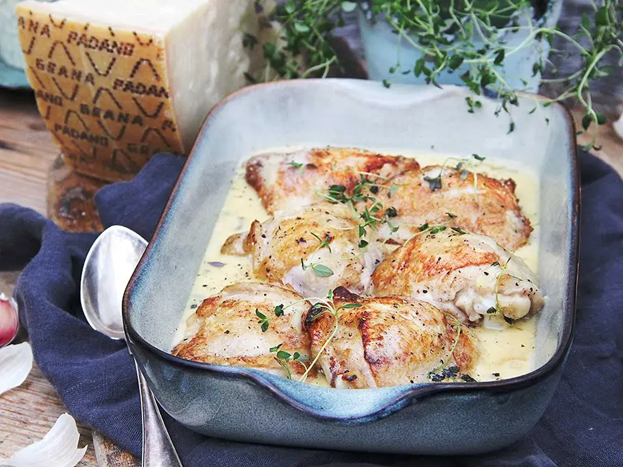 Kycklinglår i krämig sås, toppad med färska örter. En bit Grana Padano-ost syns i bakgrunden, redo att rivas över.