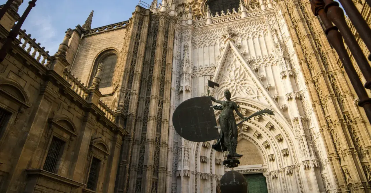 Estatua frente a la imponente fachada de la catedral de Sevilla, con detalles góticos y una escultura que evoca la navegación.