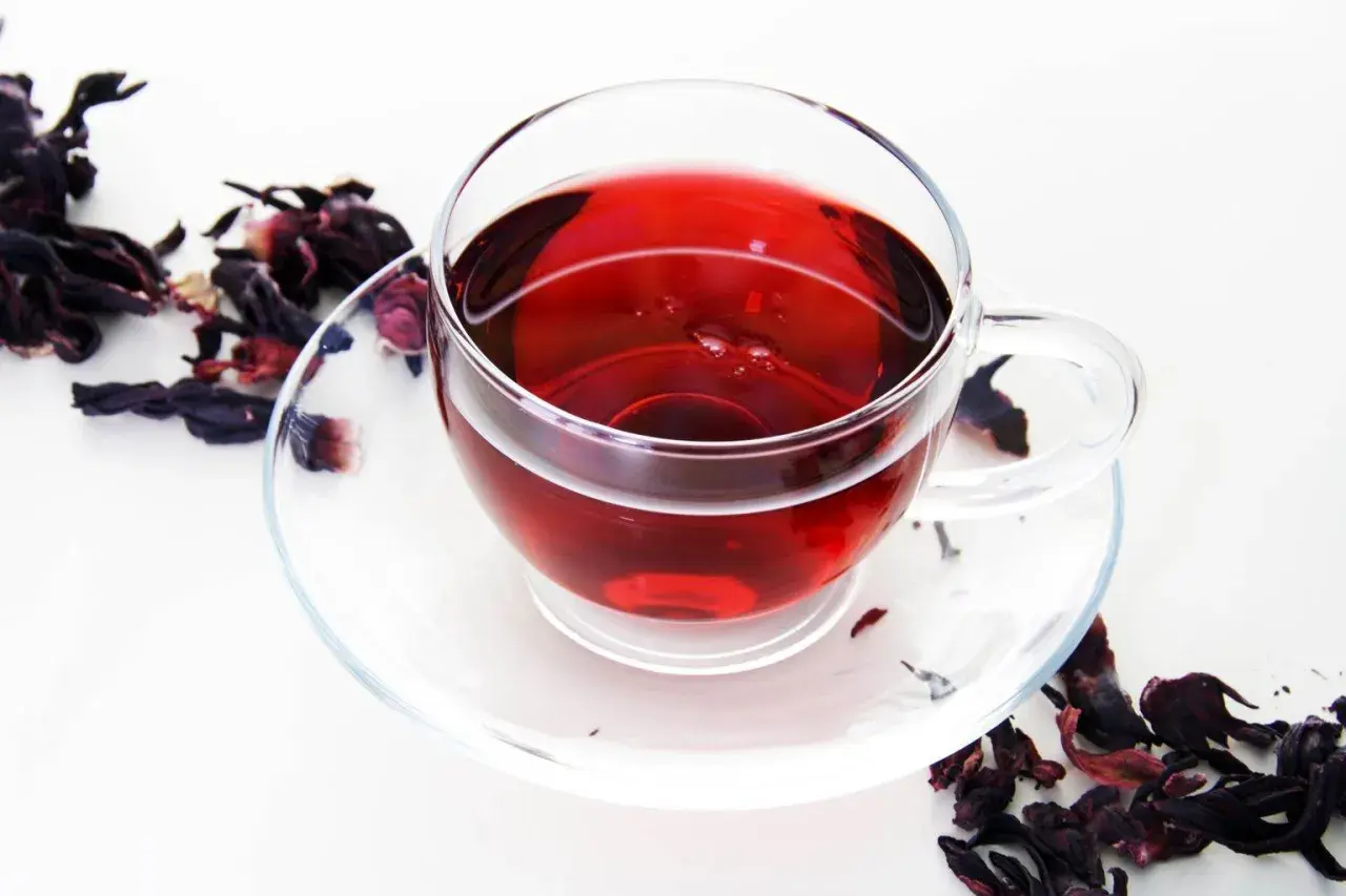 Szklana filiżanka z czerwonym naparem, obok suszone płatki hibiskusa. Idealny napój na odchudzanie.