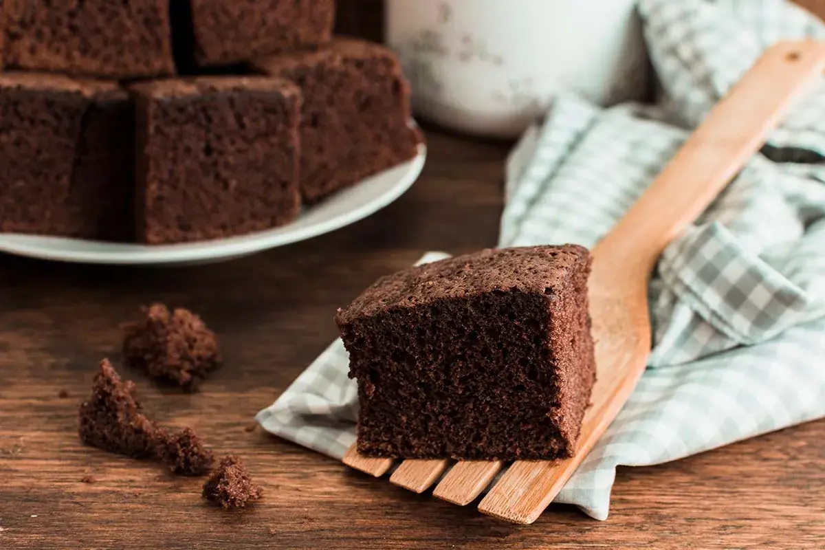 Un trozo de bizcocho de chocolate esponjoso servido en una espátula de madera, con más trozos apilados en un plato al fondo.