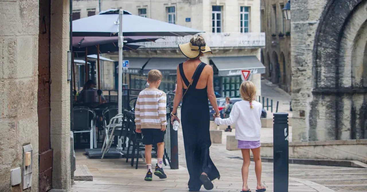 Kobieta w letniej sukience i kapeluszu prowadzi dwójkę dzieci, chłopca i dziewczynkę, ulicą. To francuskie dziecko, które zwiedza miasto.