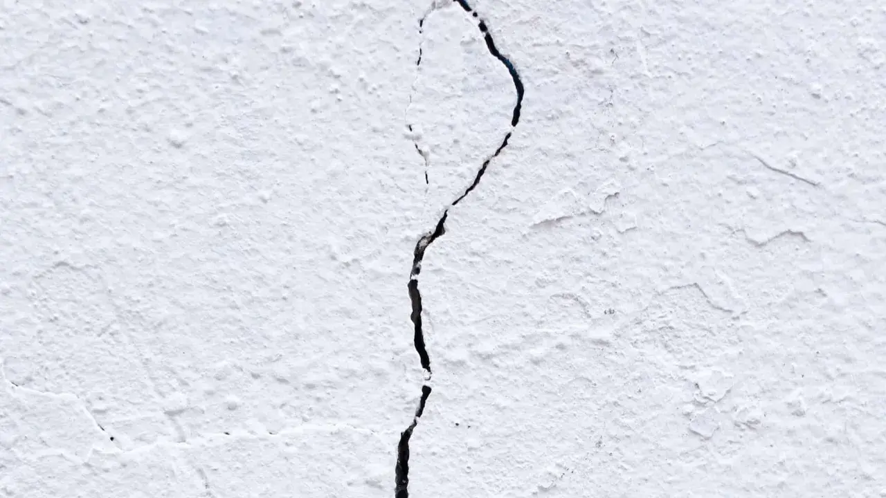 Una pared blanca con textura rugosa muestra grietas por dilatación, una línea oscura que se abre paso a través de la superficie.