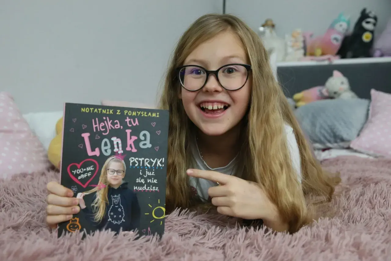 Hejka, tu Lenka! Dziewczynka z okularami trzyma notatnik z napisem "Hejka, tu Lenka".