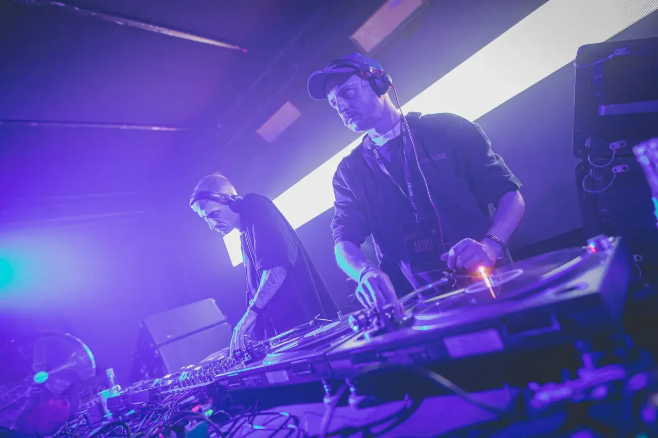Dwóch DJ-ów w słuchawkach tworzy muzykę techno. Jeden z nich ma na szyi identyfikator "ARTIST".