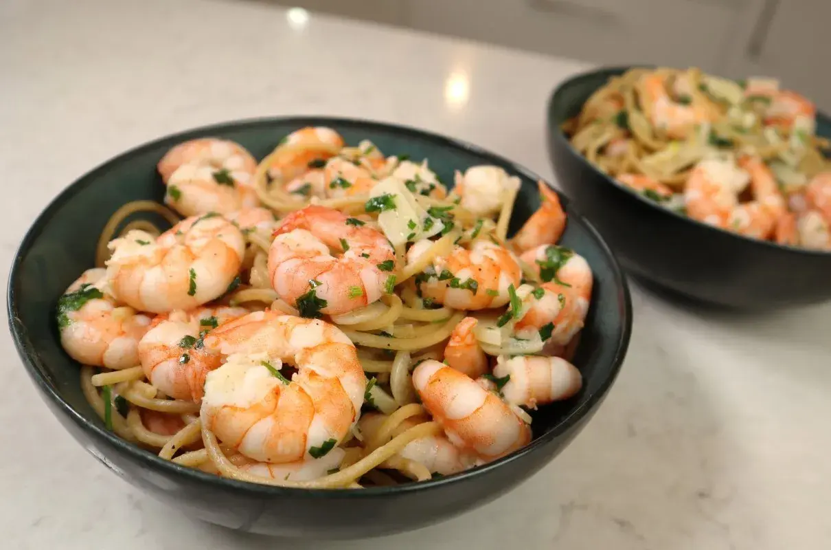 Pyszny makaron z krewetkami, posypany świeżą natką pietruszki i parmezanem, podany w ciemnych miseczkach.