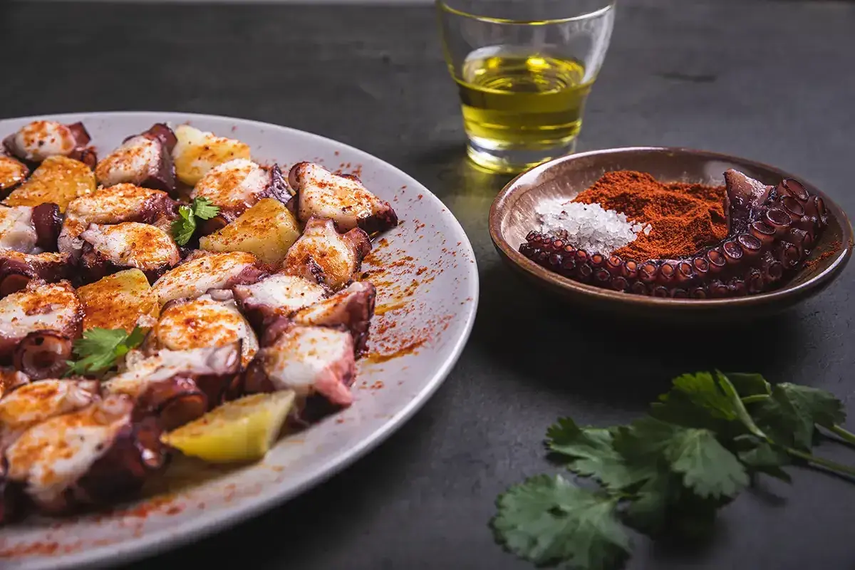 Un plat de poulpe à la gallega, garni de pommes de terre et d'épices, avec un petit bol de paprika et de sel, et un verre d'huile d'olive.