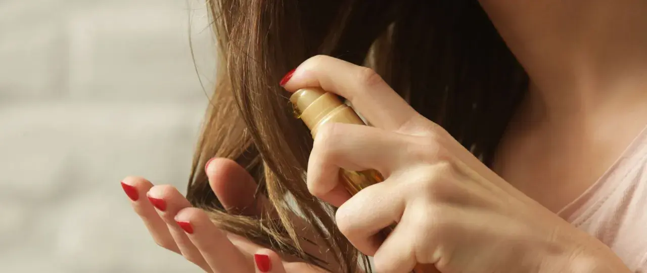Eine Hand mit roten Nägeln sprüht ein goldenes Öl auf trockene Haare, um sie zu pflegen.