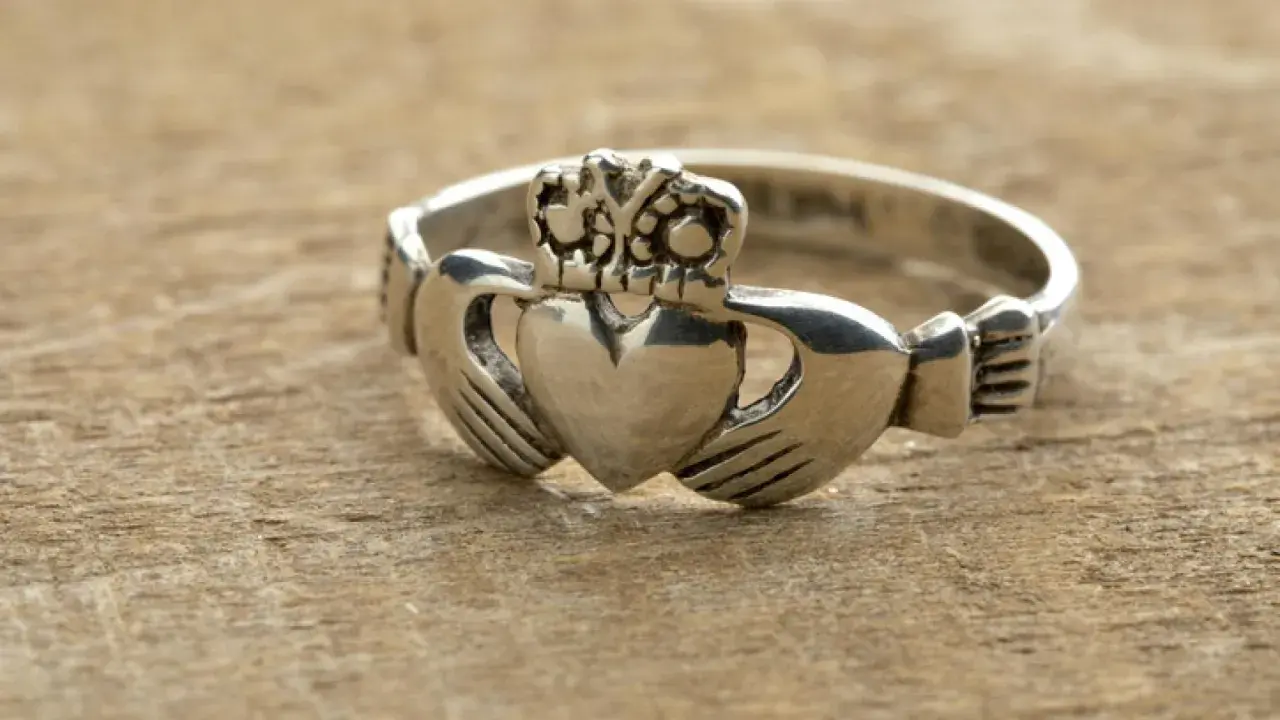Anillo Claddagh de color oro blanco, con corazón, manos y corona, sobre madera.