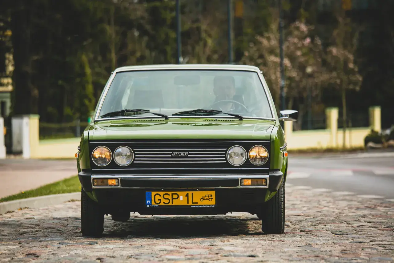 Zielony Fiat 131 Mirafiori z lat 70. na brukowanej ulicy, z widocznym kierowcą za kierownicą.