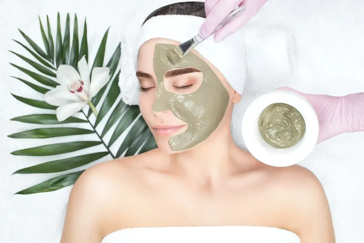 Una donna riceve un trattamento viso con maschera all'argilla verde, applicata con un pennello. Accanto, una foglia di palma e un fiore bianco.