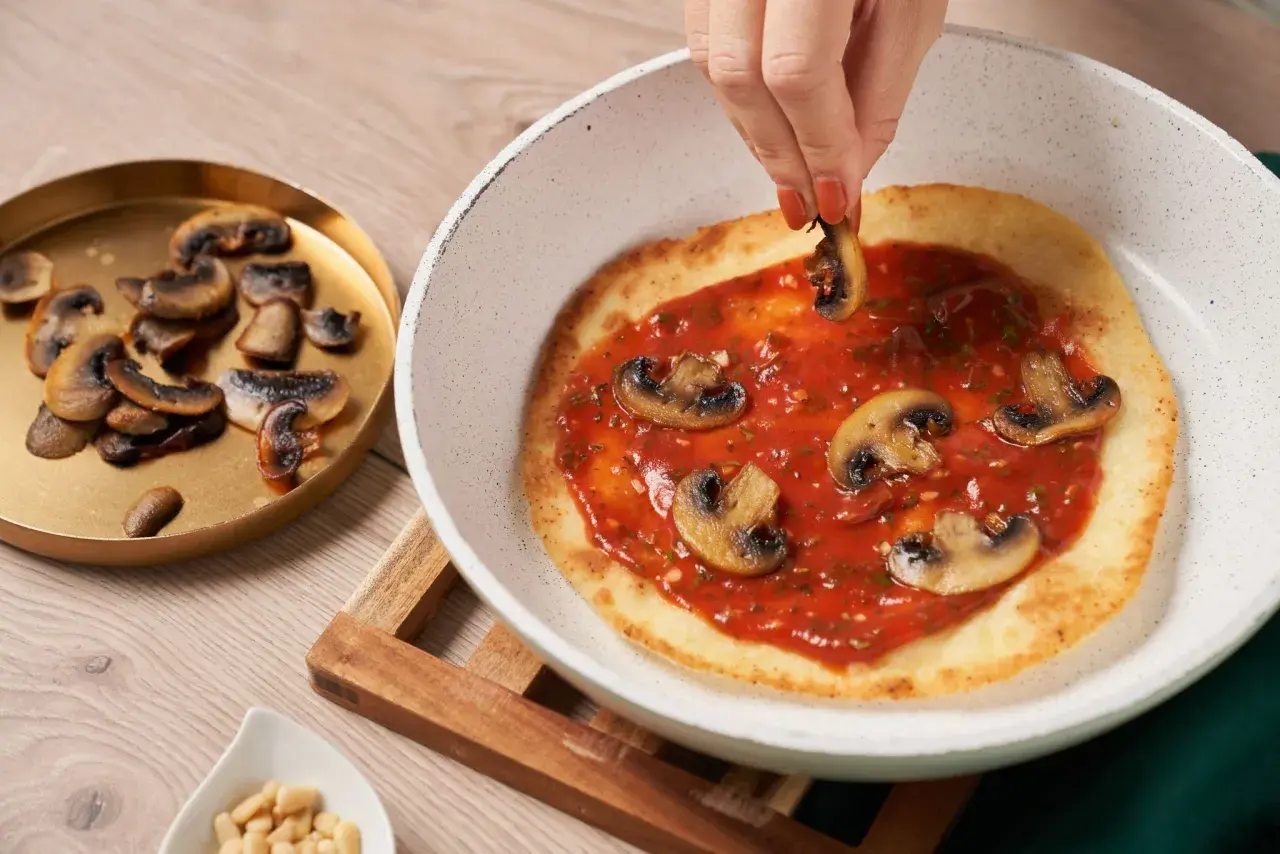 En hand lägger champinjoner på en pizza i stekpanna med tomatsås.