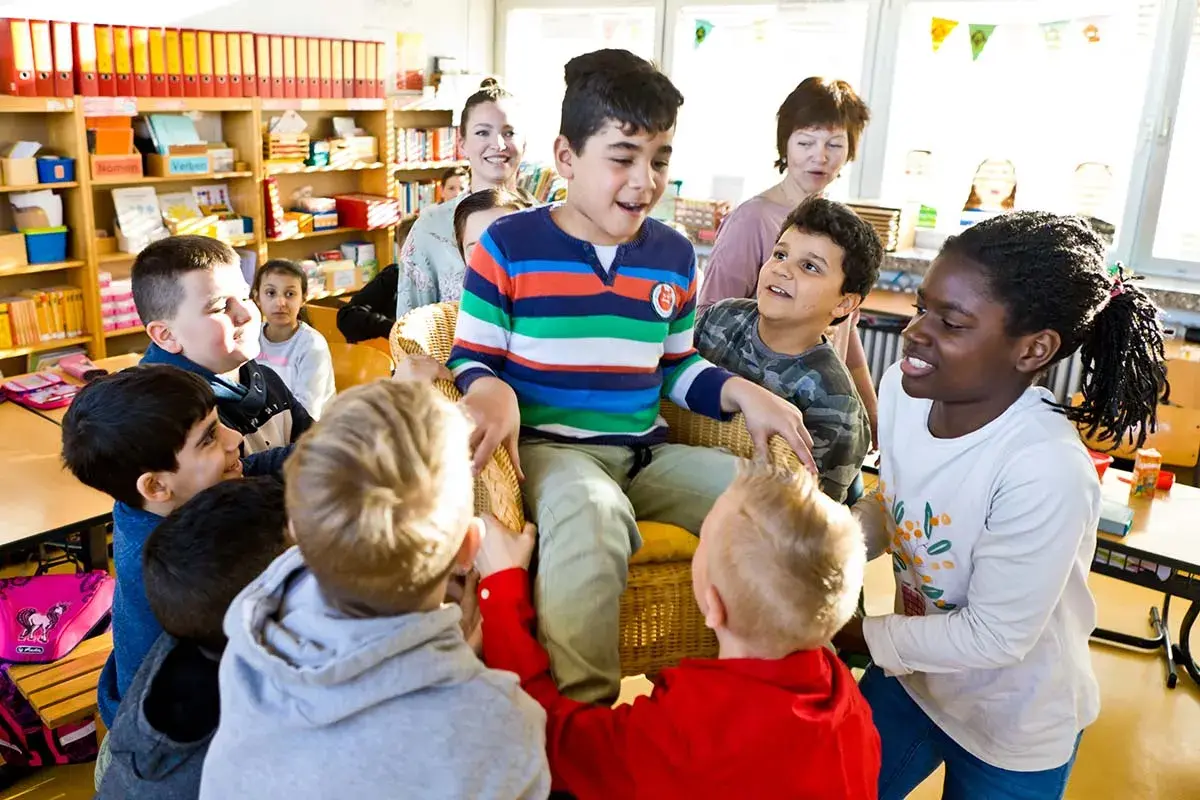 Kinder umringen einen Jungen im Stuhl. Ein lebendiges Bild vom **Unterricht Grundschule**, wo Lernen Spaß macht.