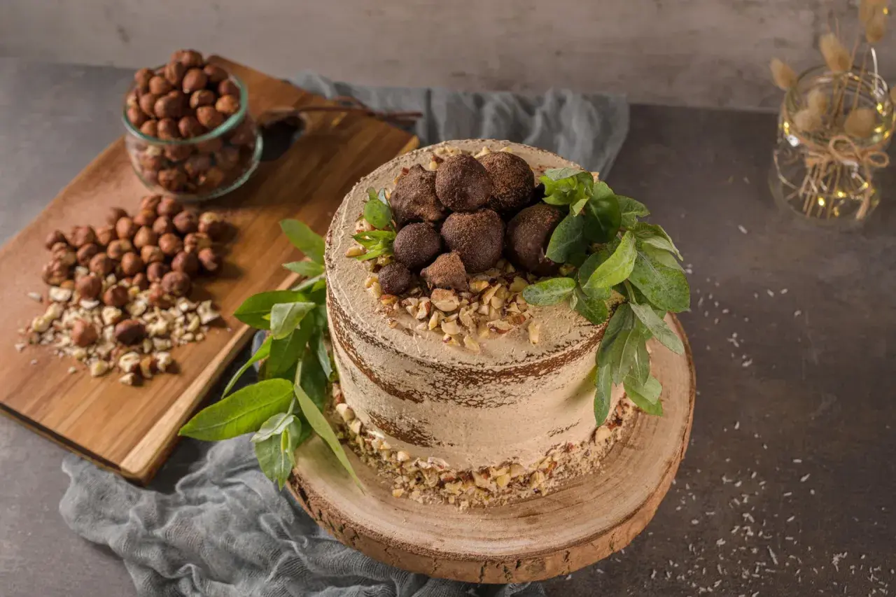 Kuchen ohne backen, verziert mit Schokoladentrüffeln, gehackten Haselnüssen und grünen Blättern. Daneben ein Glas mit Haselnüssen.