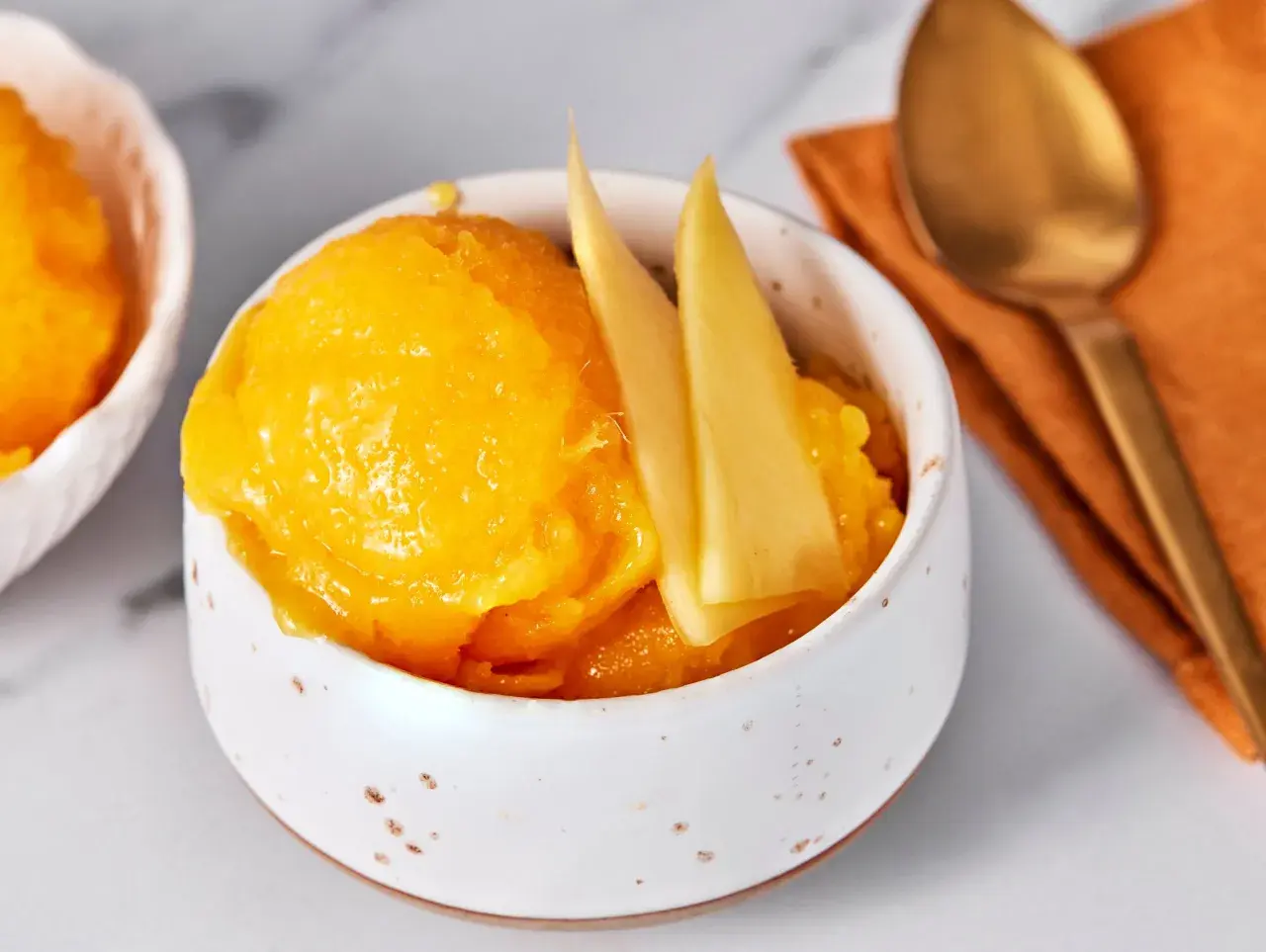 Erfrischendes Mango Sorbet in einer Schale, garniert mit Mangostücken. Daneben ein Löffel auf einem orangen Tuch.
