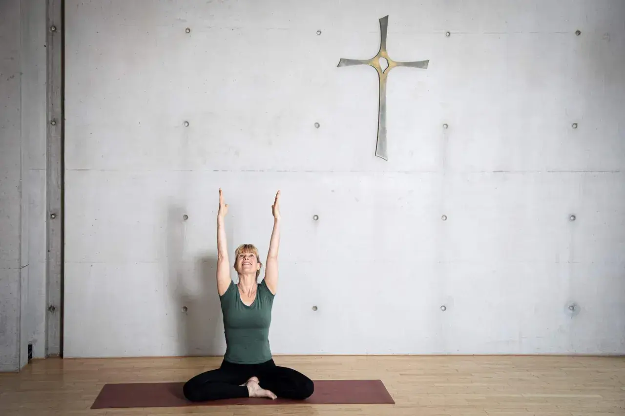Frau in Yoga-Pose mit erhobenen Armen vor einem Kreuz an einer Betonwand. Eine harmonische Verbindung von Yoga und Christentum.