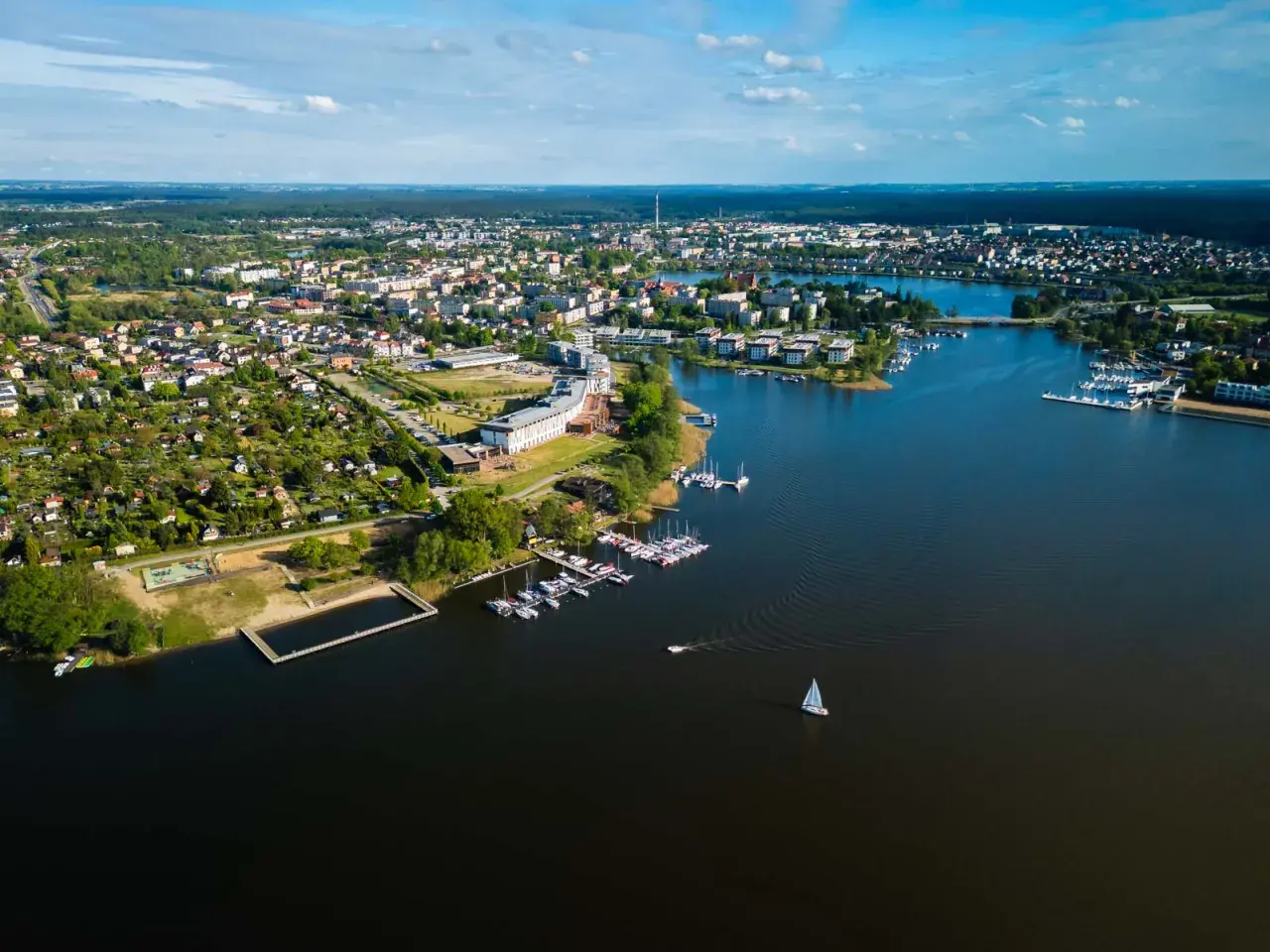 Malownicze miasto nad Jeziorakiem, z mariną pełną żaglówek i łodzi, otoczone zielenią i zabudową mieszkalną.