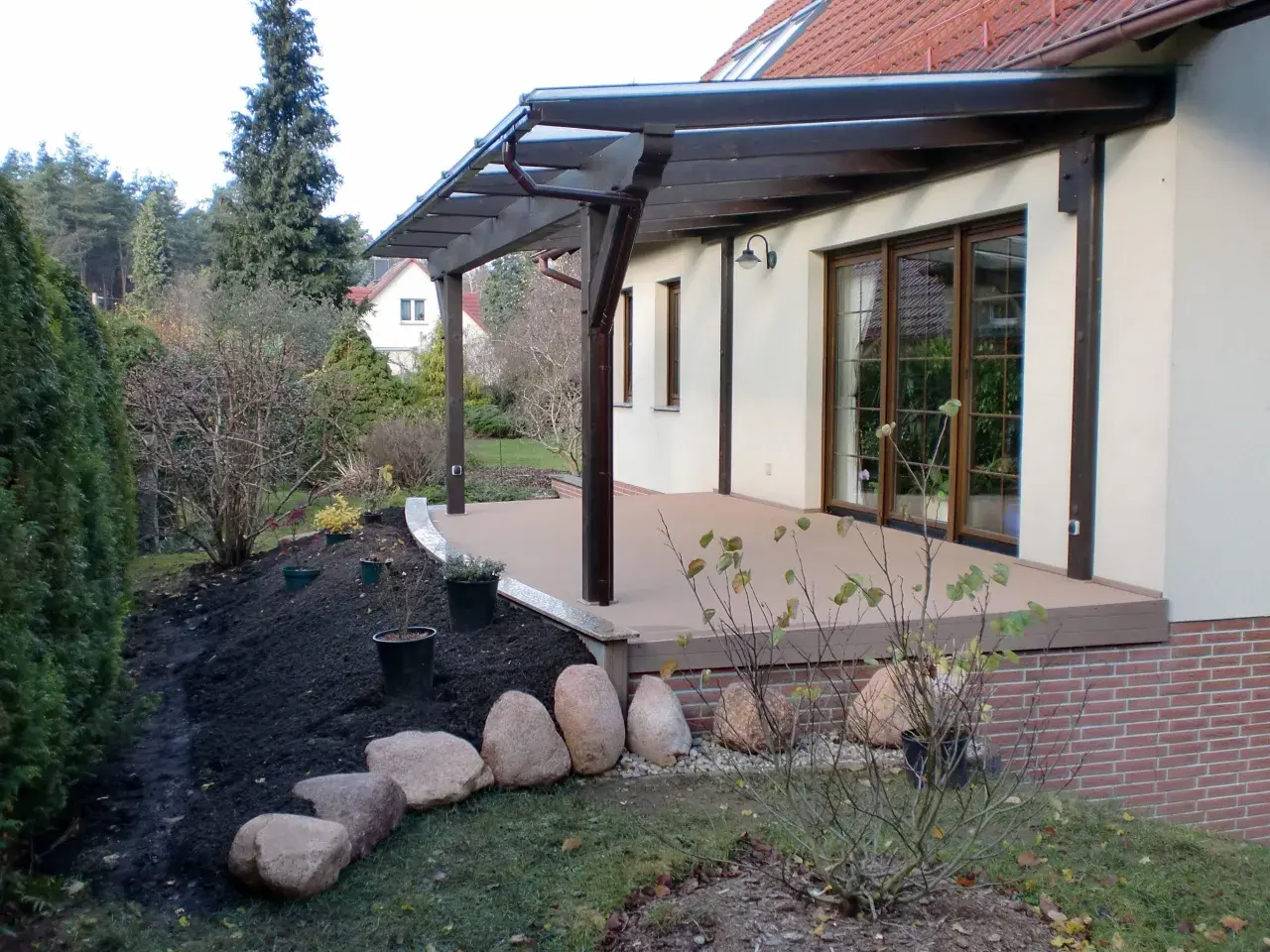 Terrasse sanieren: Neue Holzterrasse mit Überdachung, daneben frisch aufgeschüttete Erde und Steine für die Gartengestaltung.