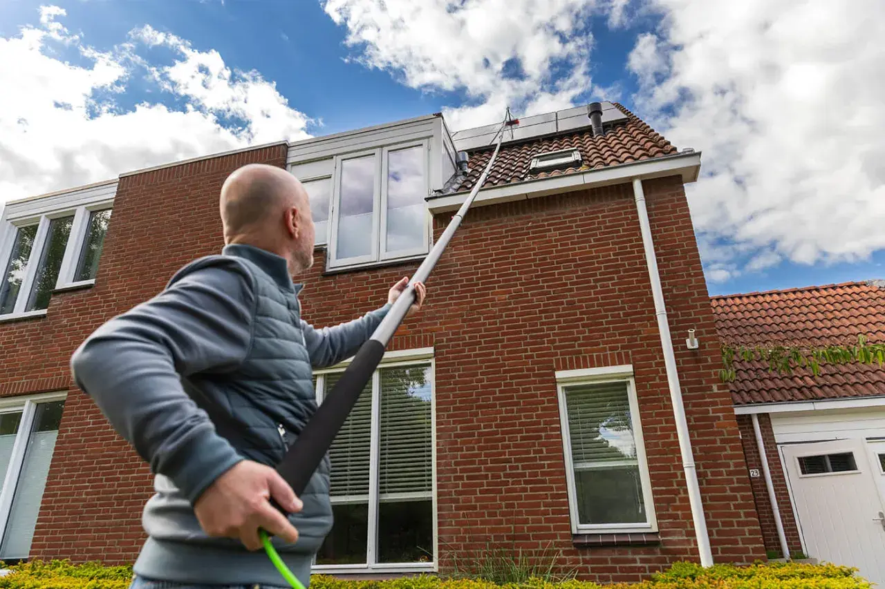 Hombre con pértiga extendida para limpiar tejas de una casa de ladrillo rojo bajo un cielo azul con nubes.