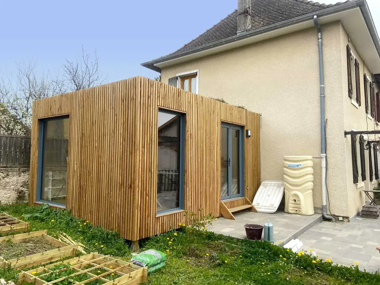 Une extension en bois moderne s'intègre à une maison existante, avec de grandes fenêtres et une finition en lattes de bois.