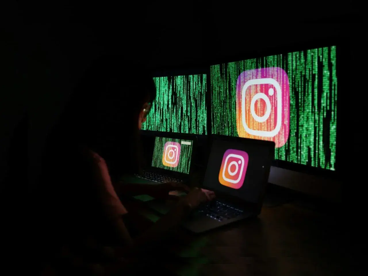 Osoba pracuje przy komputerze, na ekranach widnieje logo Instagrama i zielony kod. Szuka informacji, jak odzyskać zhakowane konto na Instagramie.