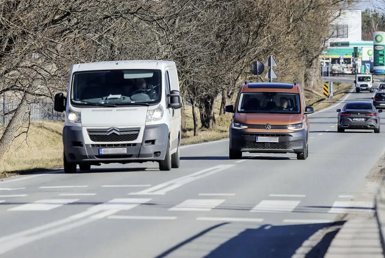 Biała furgonetka wyprzedza na przejściu dla pieszych pomarańczowego vana. W tle stacja benzynowa.