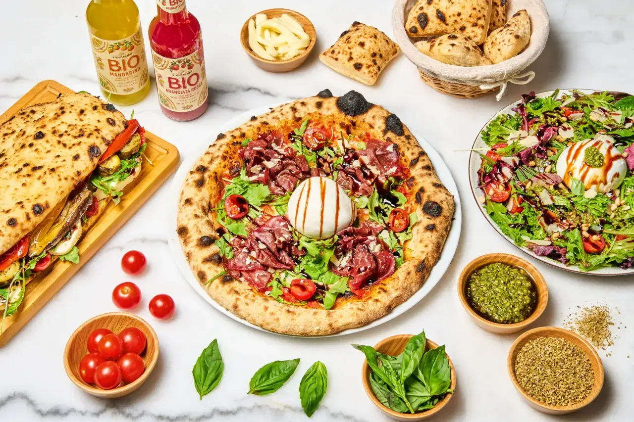 Une pizza diavola appétissante, garnie de charcuterie, burrata et roquette, accompagnée d'un sandwich, de salade et de boissons bio.