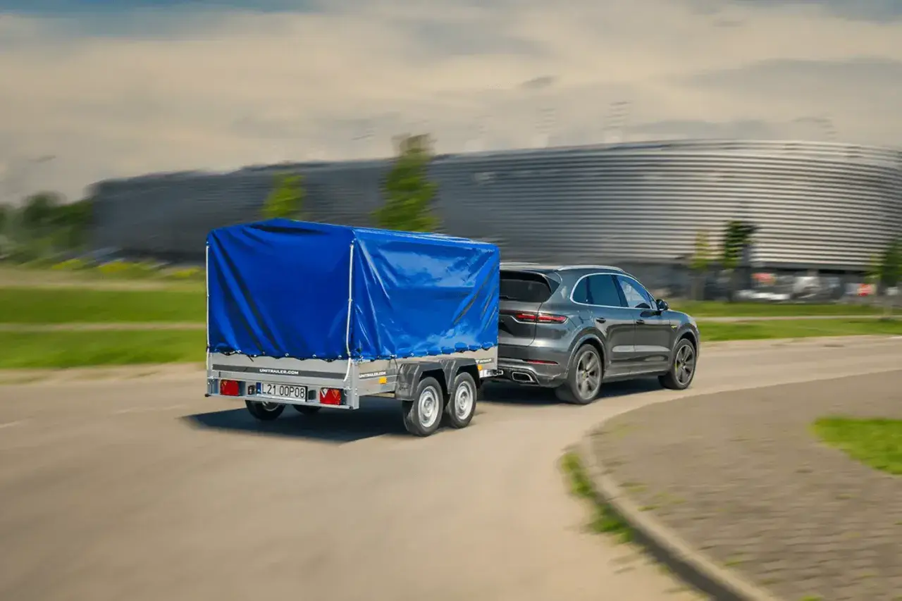 Szary SUV ciągnie dwuosiową przyczepę pod niebieską plandeką. Czy można ciągnąć przyczepę dwuosiową na kat. B?