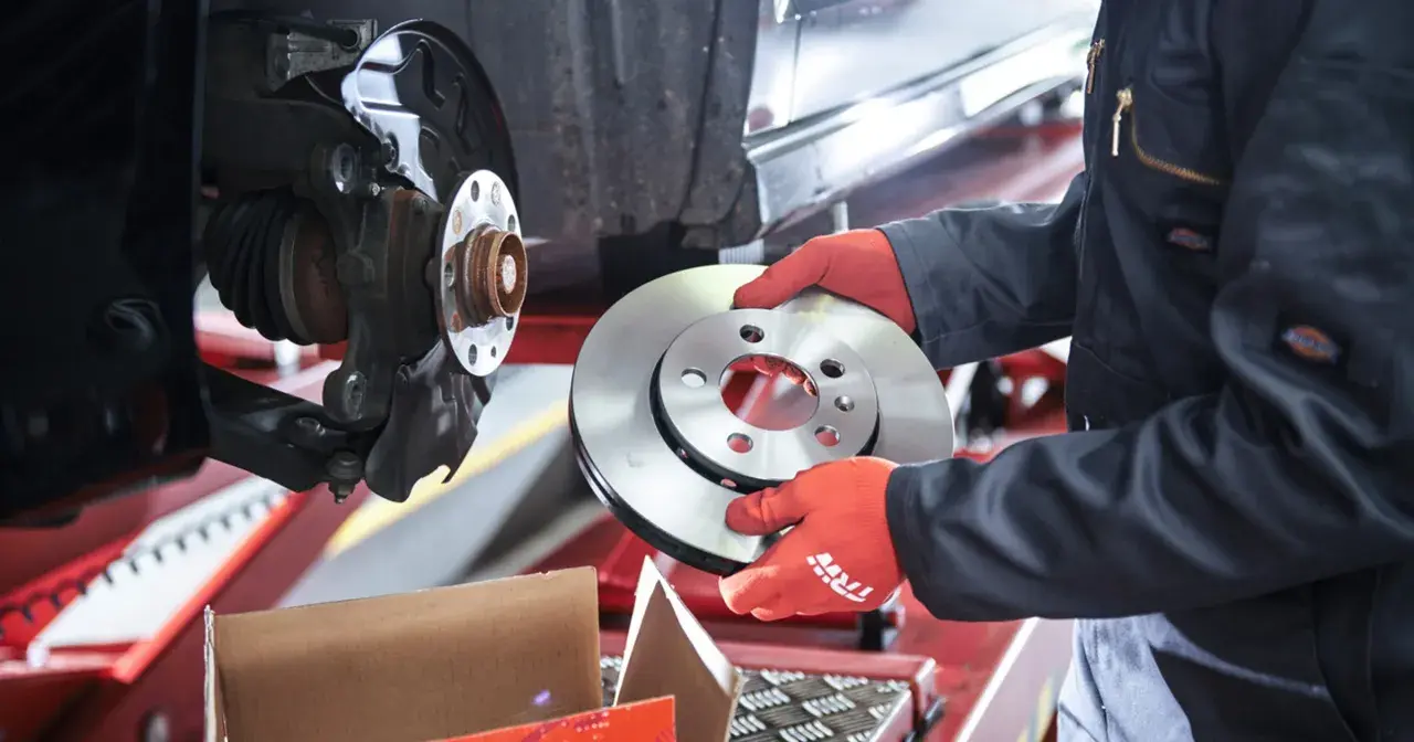 Mechanik w czerwonych rękawiczkach trzyma nową tarczę hamulcową obok zamontowanej tarczy.