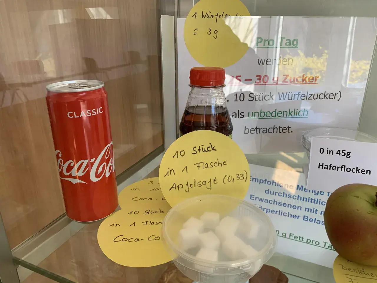 Eine Cola-Dose neben Würfelzucker. Die Frage "wie viel gramm zucker hat cola" wird durch Notizen beantwortet: 10 Stück Würfelzucker pro Flasche.