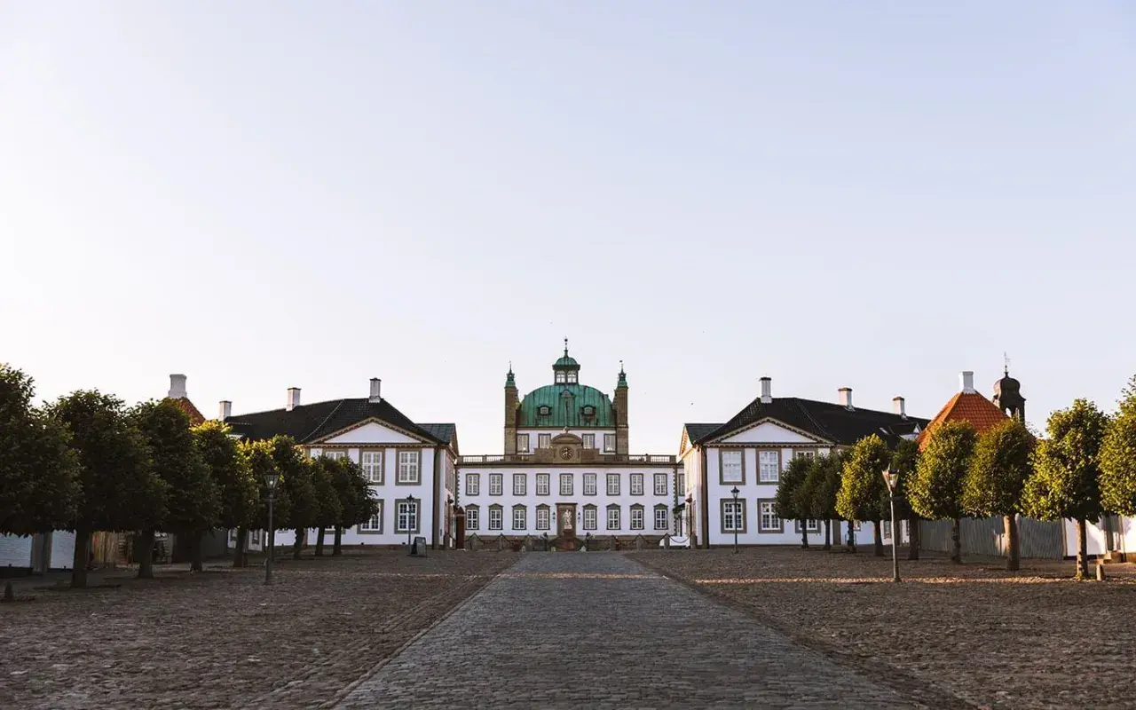 Frederiksborgs slott med sin gröna kupol reser sig majestätiskt mot en klar himmel. En stenlagd väg leder fram till den imponerande byggnaden, flankerad av träd.