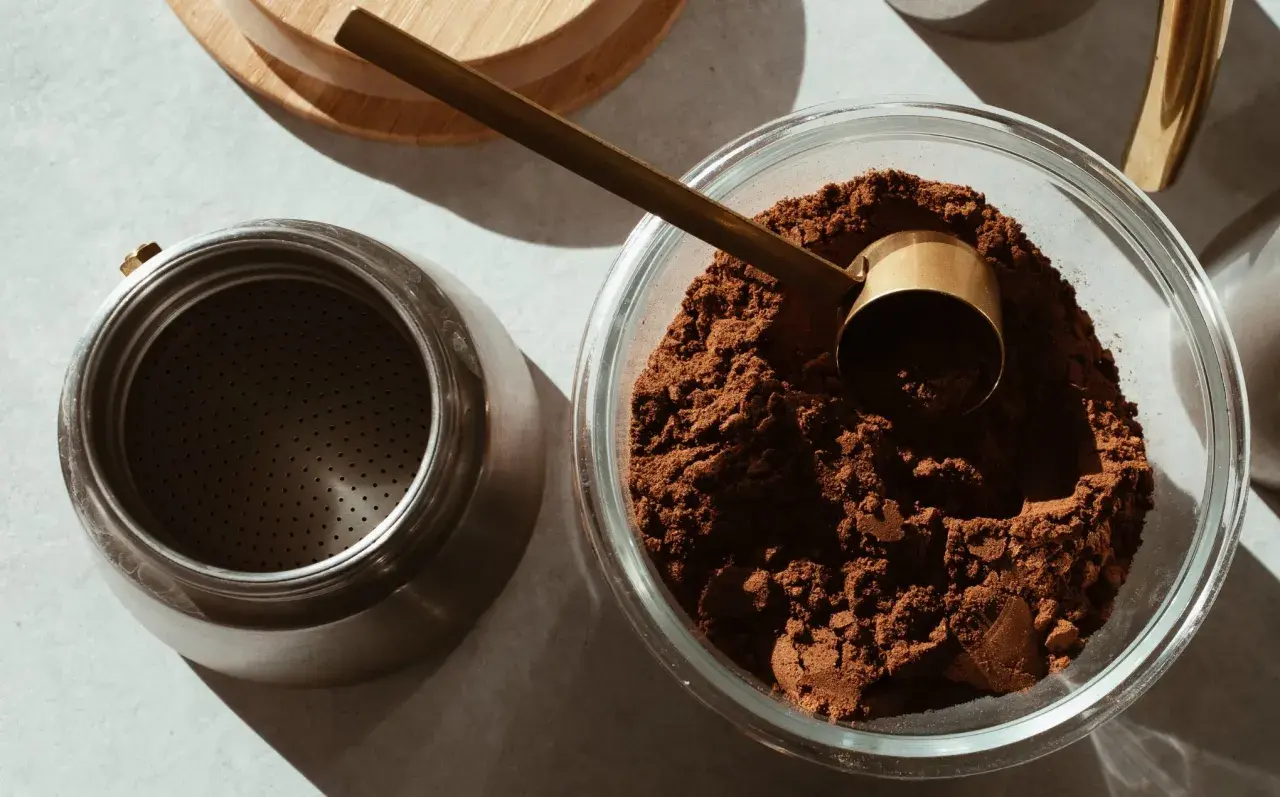 Caffè macinato in una ciotola di vetro, pronto per essere conservato. Un misurino dorato e una moka completano la scena, suggerendo come conservare il caffè al meglio.