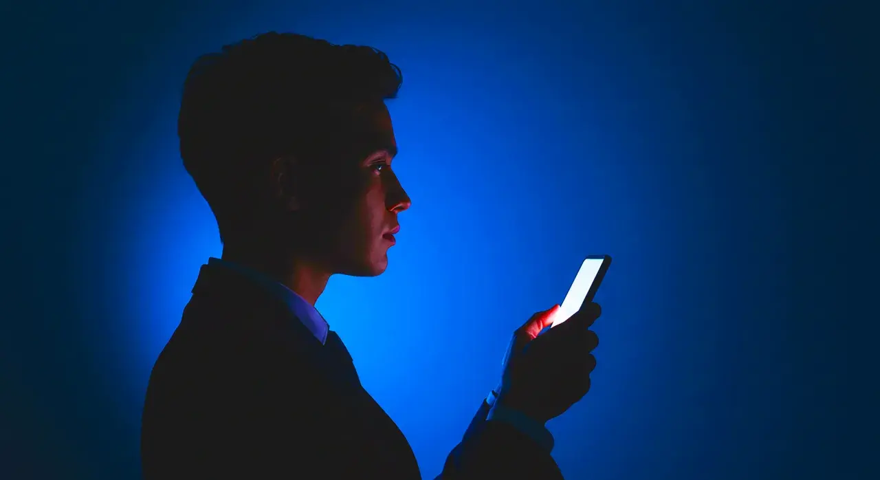 Mężczyzna w garniturze patrzy na jasny ekran telefonu. Dba o ochronę wzroku w telefonie, używając go w ciemności.