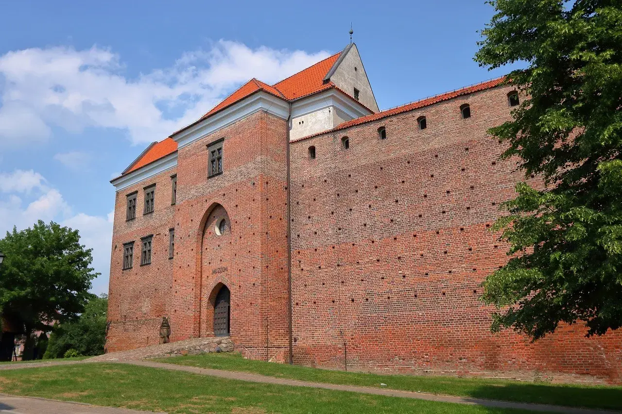 Zamek w Łęczycy, ceglana twierdza z czerwonej cegły, z charakterystycznym łukowym wejściem i oknami, otoczona zielenią.