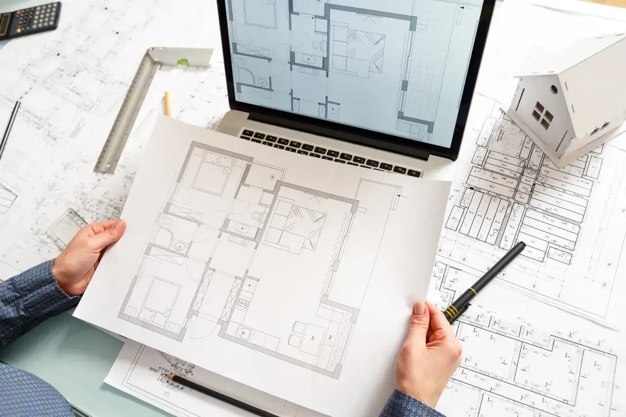 Architekt analizuje plany mieszkania, które staną się jego pierwszymi kluczami do sukcesu. Na stole rysunki, laptop i makieta domu.