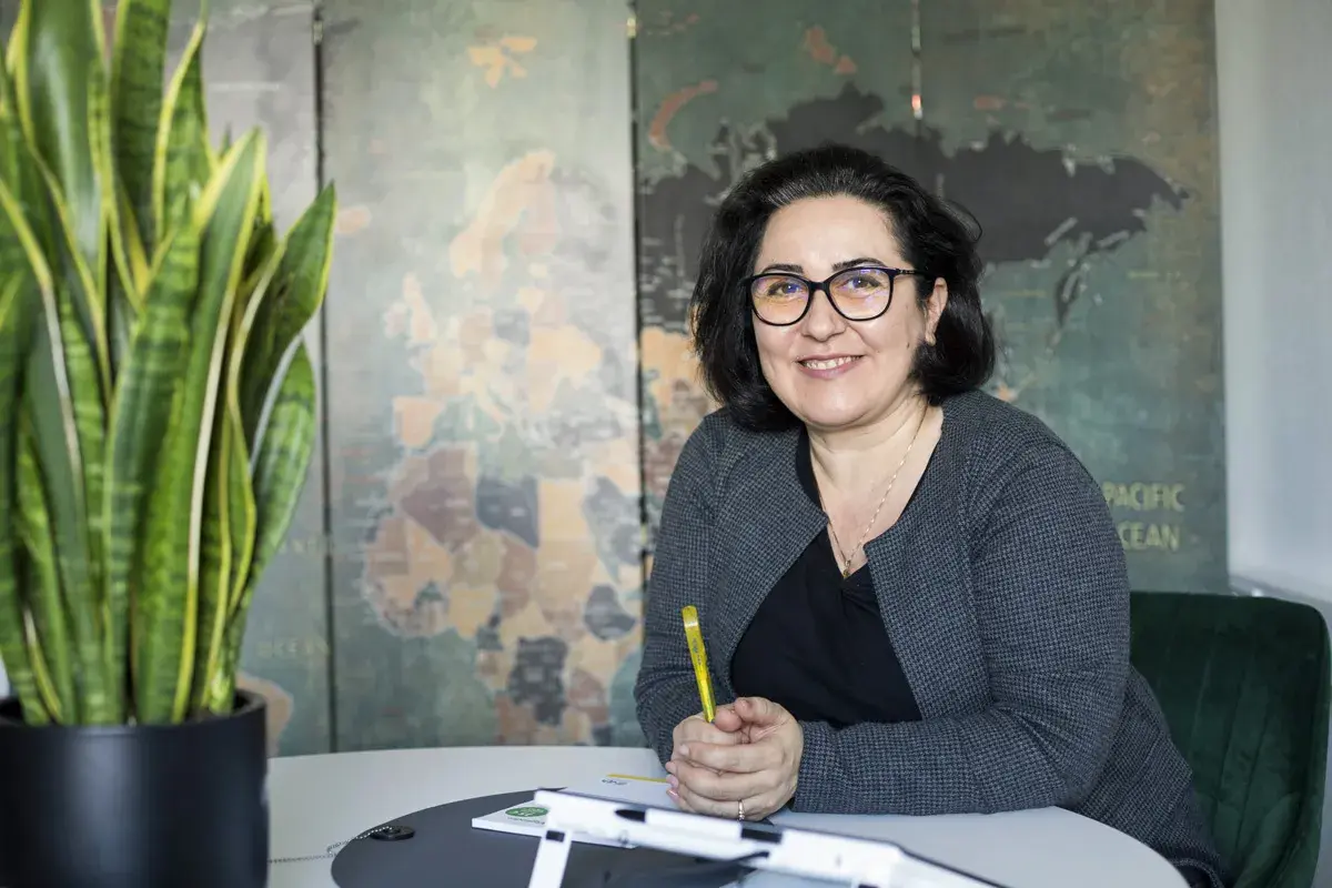 Frau mit Brille lächelt, hält Stift. Im Hintergrund Weltkarte. Sie berät zu Lohnsteuerhilfeverein Kosten.