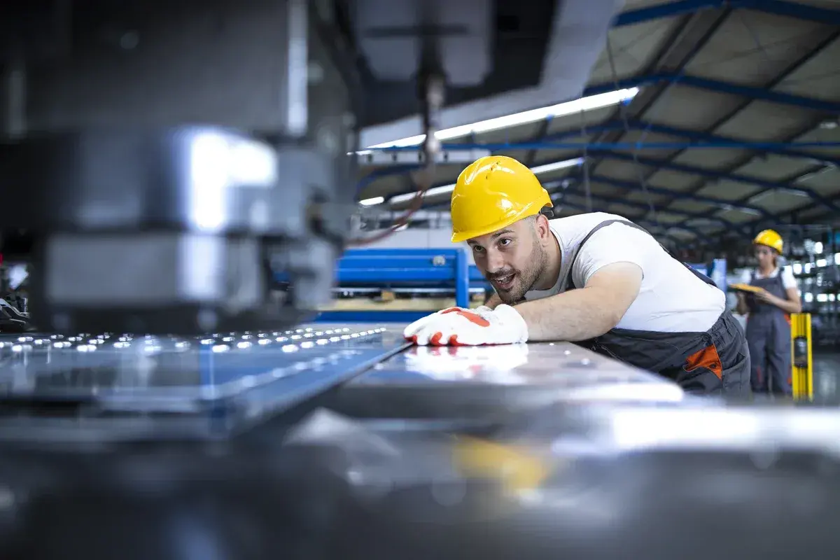 Uśmiechnięty operator CNC w żółtym kasku pochyla się nad maszyną, kontrolując proces produkcji.