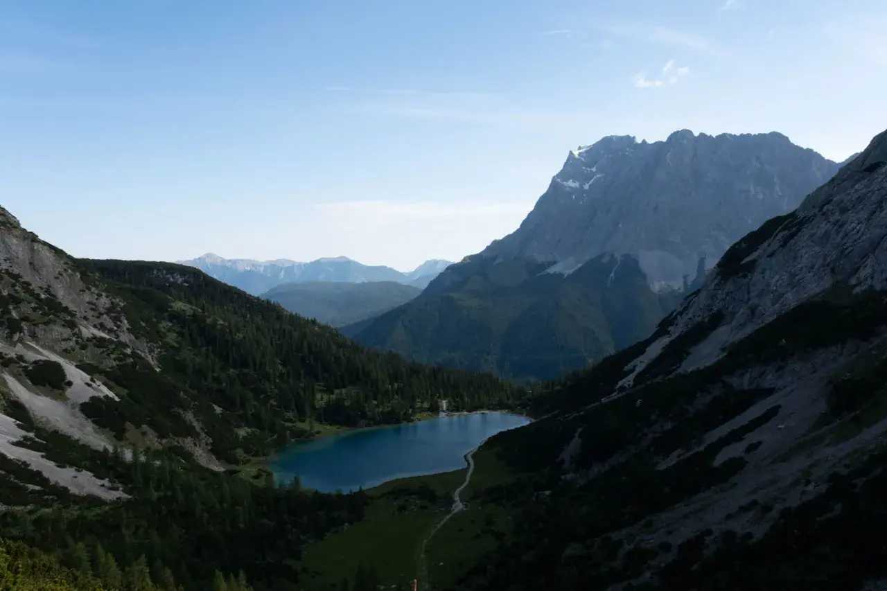 Ein Bergsee liegt eingebettet zwischen steilen Hängen, die wie ein vorderer Drachenkopf wirken. Nadelbäume bedecken die Flanken, im Hintergrund ragen weitere Gipfel auf.
