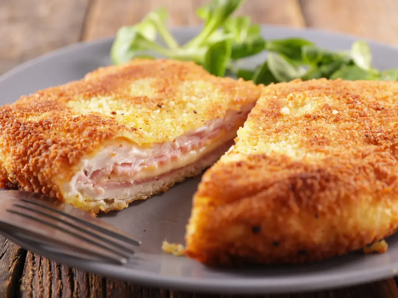 Cotoletta alla valdostana dorata e croccante, farcita con prosciutto e formaggio filante, servita su un piatto grigio con un contorno di insalata verde.