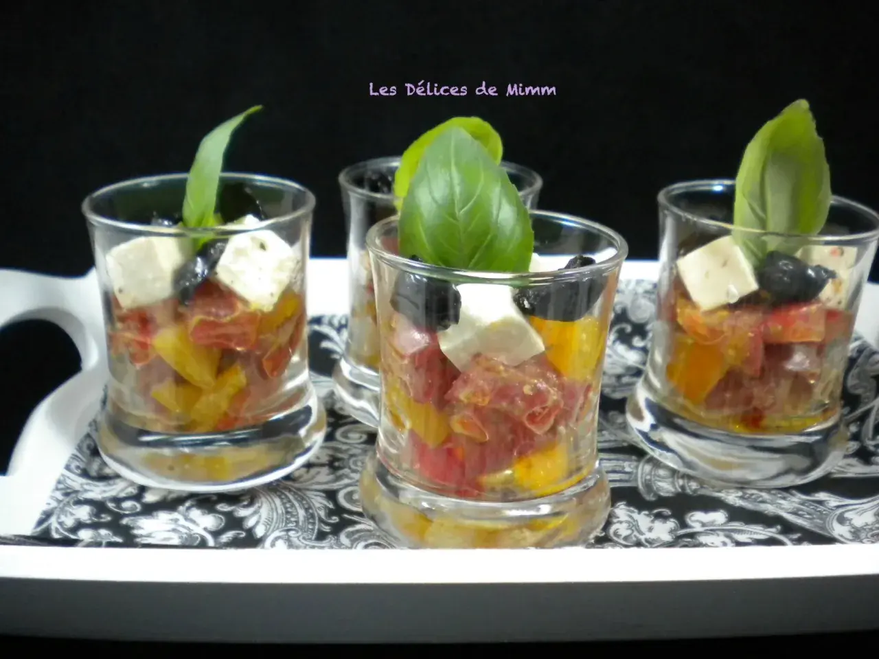 Quatre verrines apéro colorées avec tomates, olives, feta et basilic, présentées sur un plateau décoré.