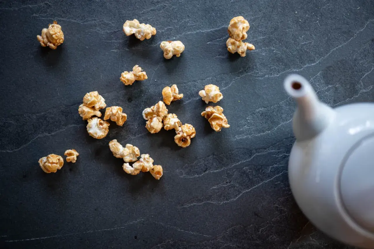Pop corn caramel éparpillé sur une surface sombre, avec une théière blanche en arrière-plan.