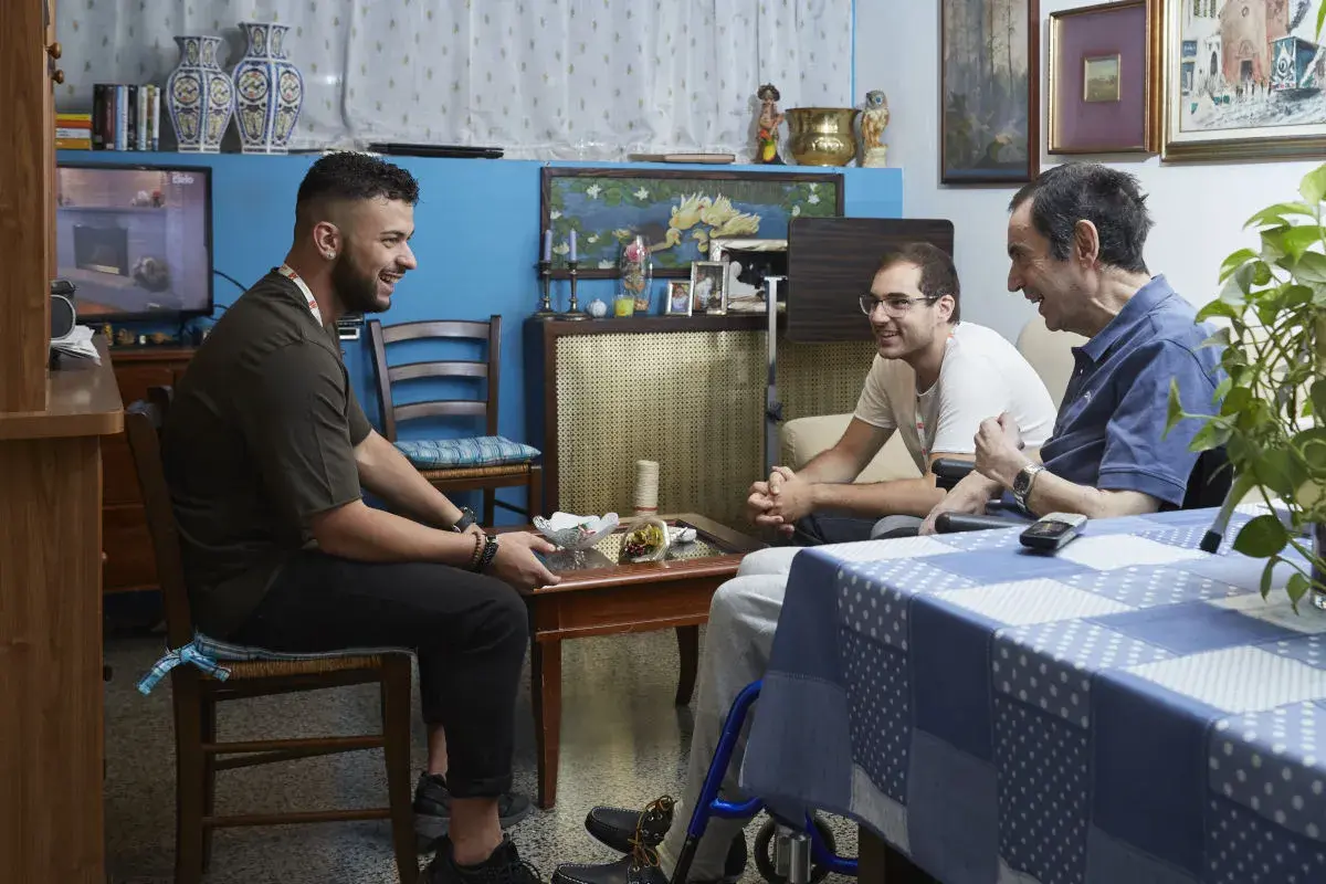 Tre uomini discutono in un salotto. Uno di loro, seduto su una sedia a rotelle, potrebbe chiedere quanto prende di pensione un dializzato.