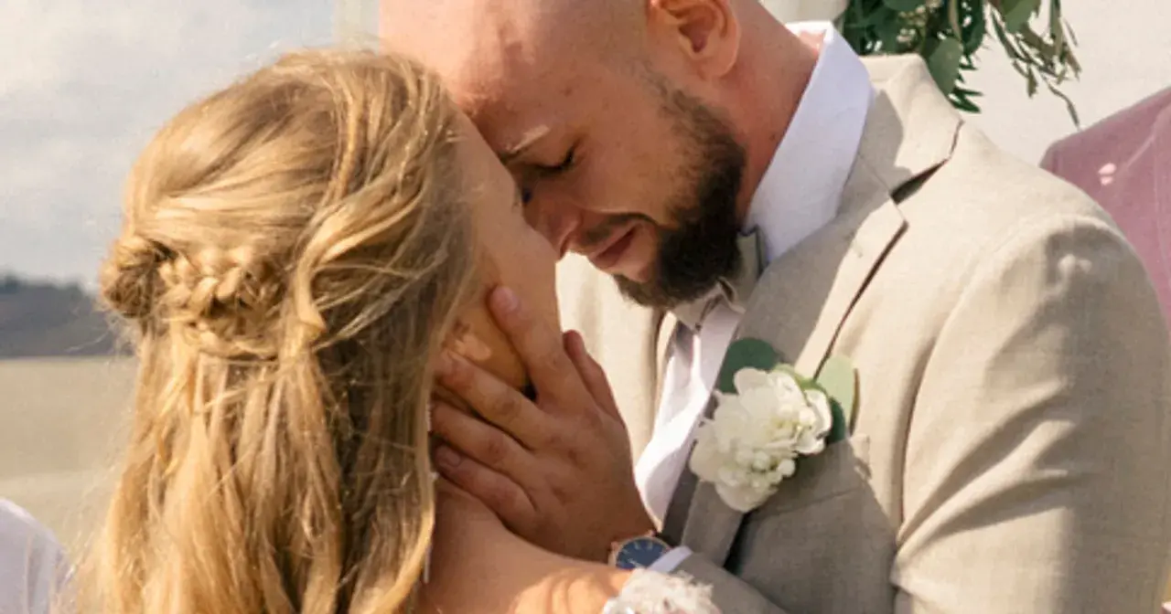 Agnieszka Kaczorowska i jej mąż w czułym uścisku podczas ślubu. Ona ma blond włosy splecione w warkocz, on w jasnej marynarce z butonierką.