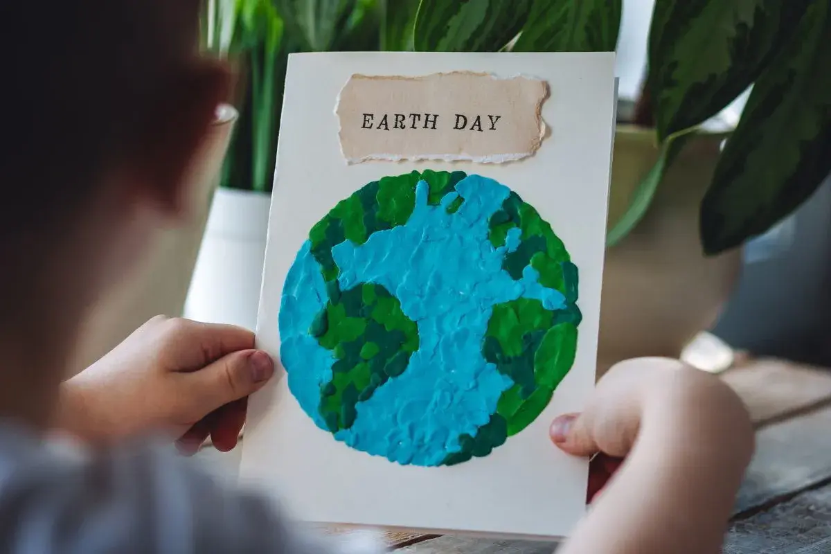 Dziecko trzyma kartkę z pracą plastyczną przedstawiającą planetę Ziemię z plasteliny. Napis "EARTH DAY" na górze.