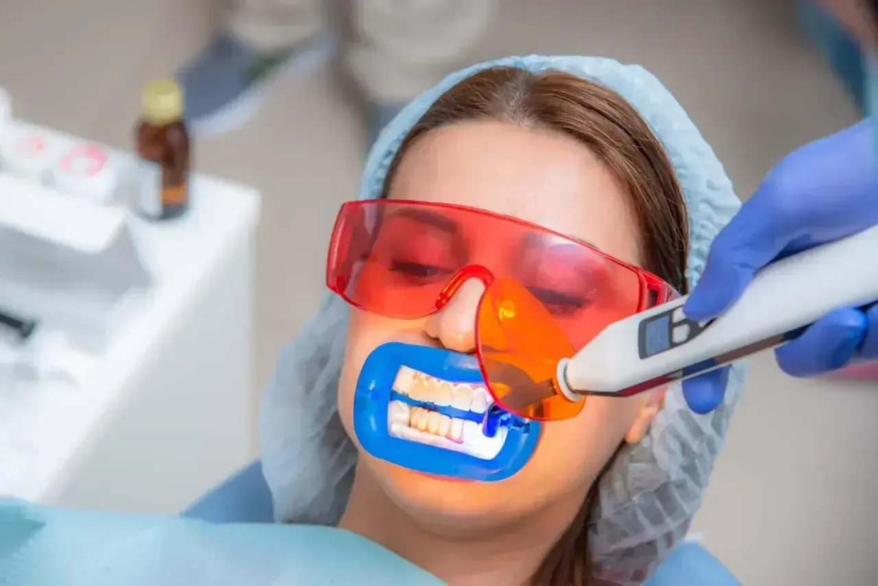 Kobieta w okularach ochronnych przechodzi zabieg lakowania zębów. Dentysta używa lampy do utwardzenia materiału.