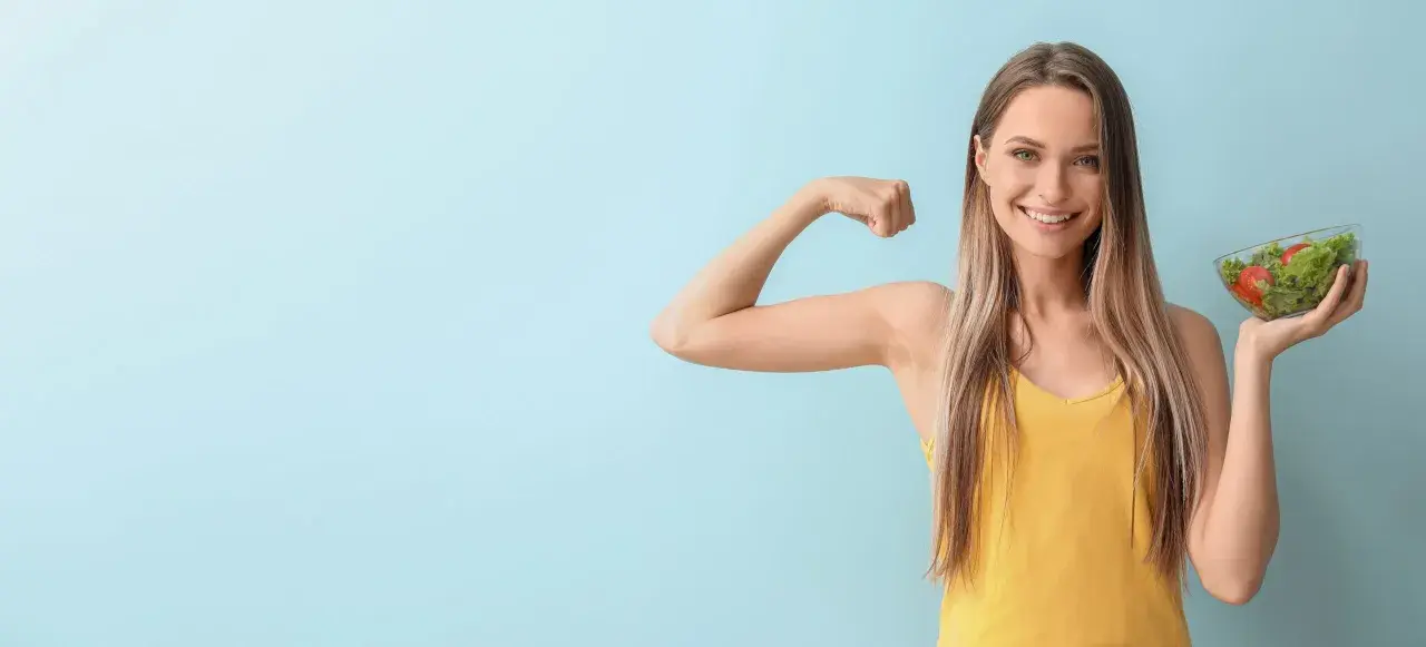 Donna sorridente mostra il bicipite e una ciotola di insalata, simbolo di una dieta per stitichezza menù settimanale salutare e benessere.