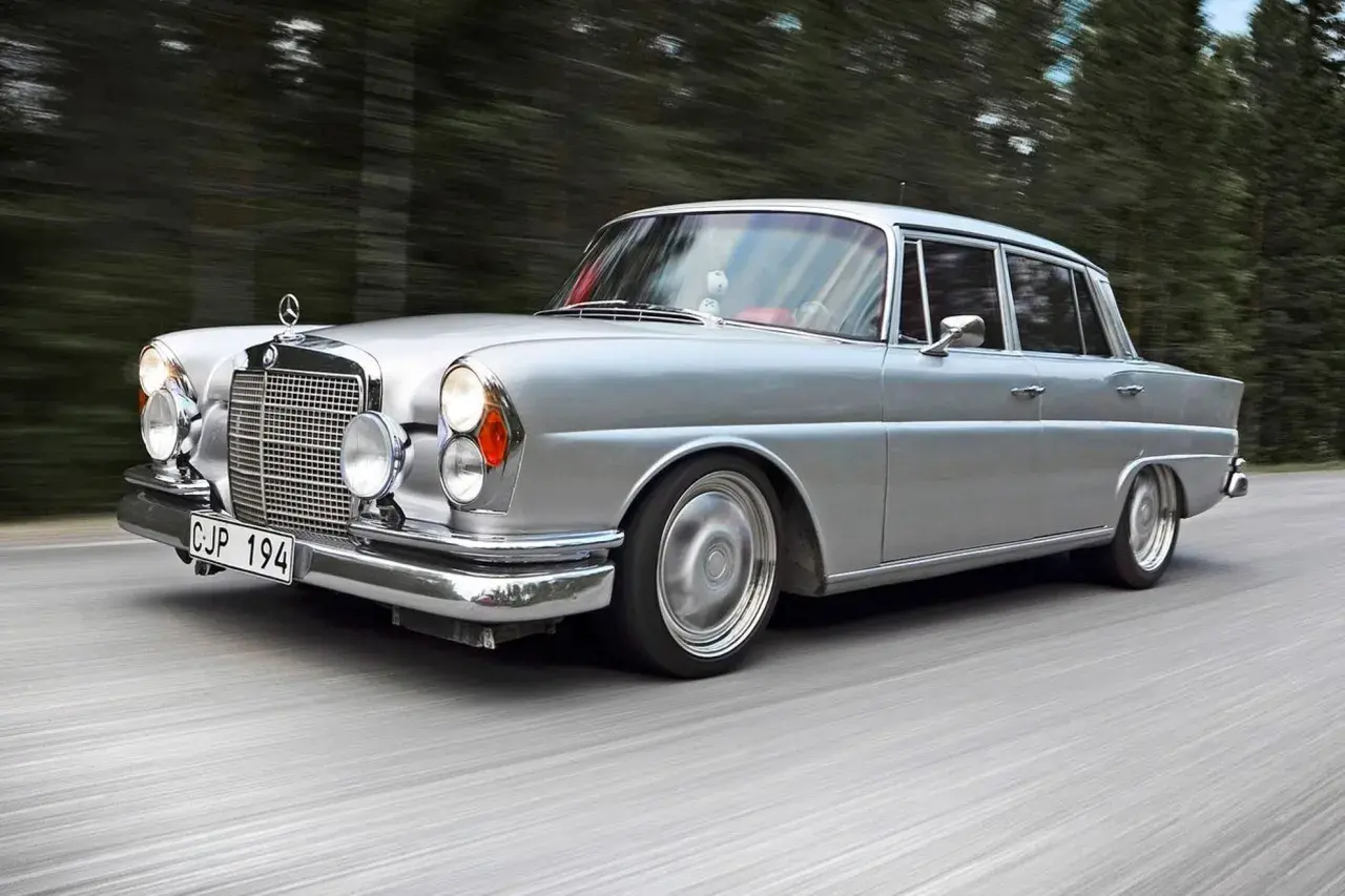 Silver Mercedes från 60-talet glider fram på en väg, omgiven av skog. Klassiska bilar som denna är tidlösa.