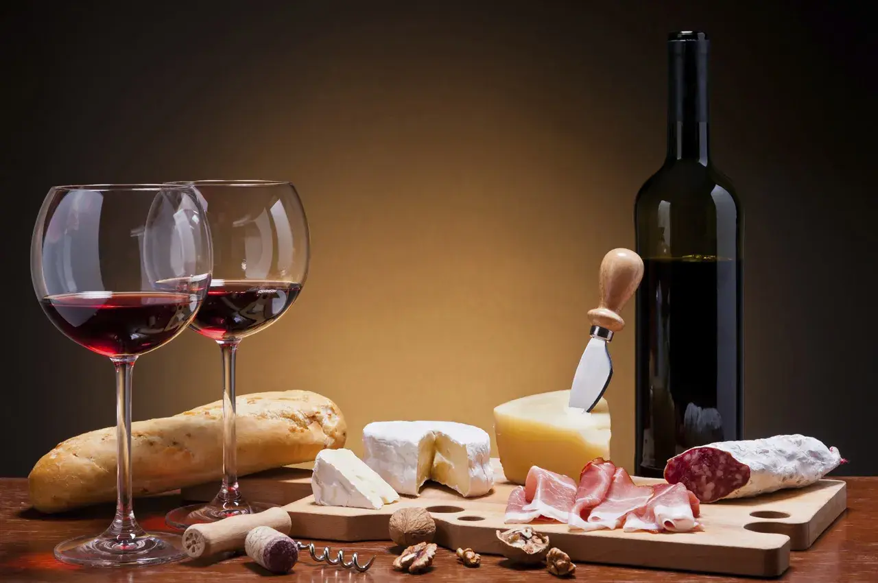 Deux verres de vin rouge, une bouteille, du pain, des fromages, de la charcuterie et des noix. Un accord parfait pour un vin pour fromage et charcuterie.