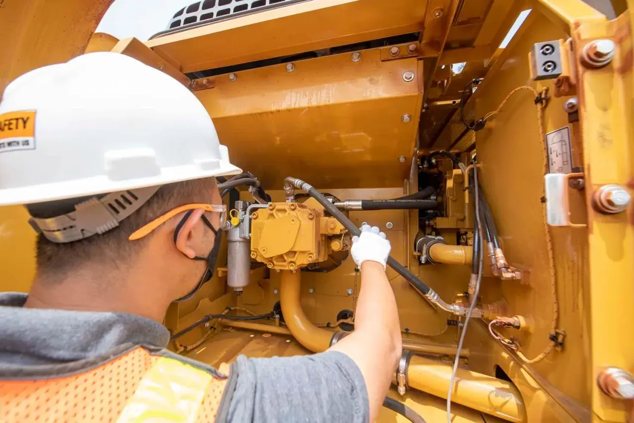 Pracownik w kasku i kamizelce konserwuje żółte urządzenia budowlane, sprawdzając przewody hydrauliczne.